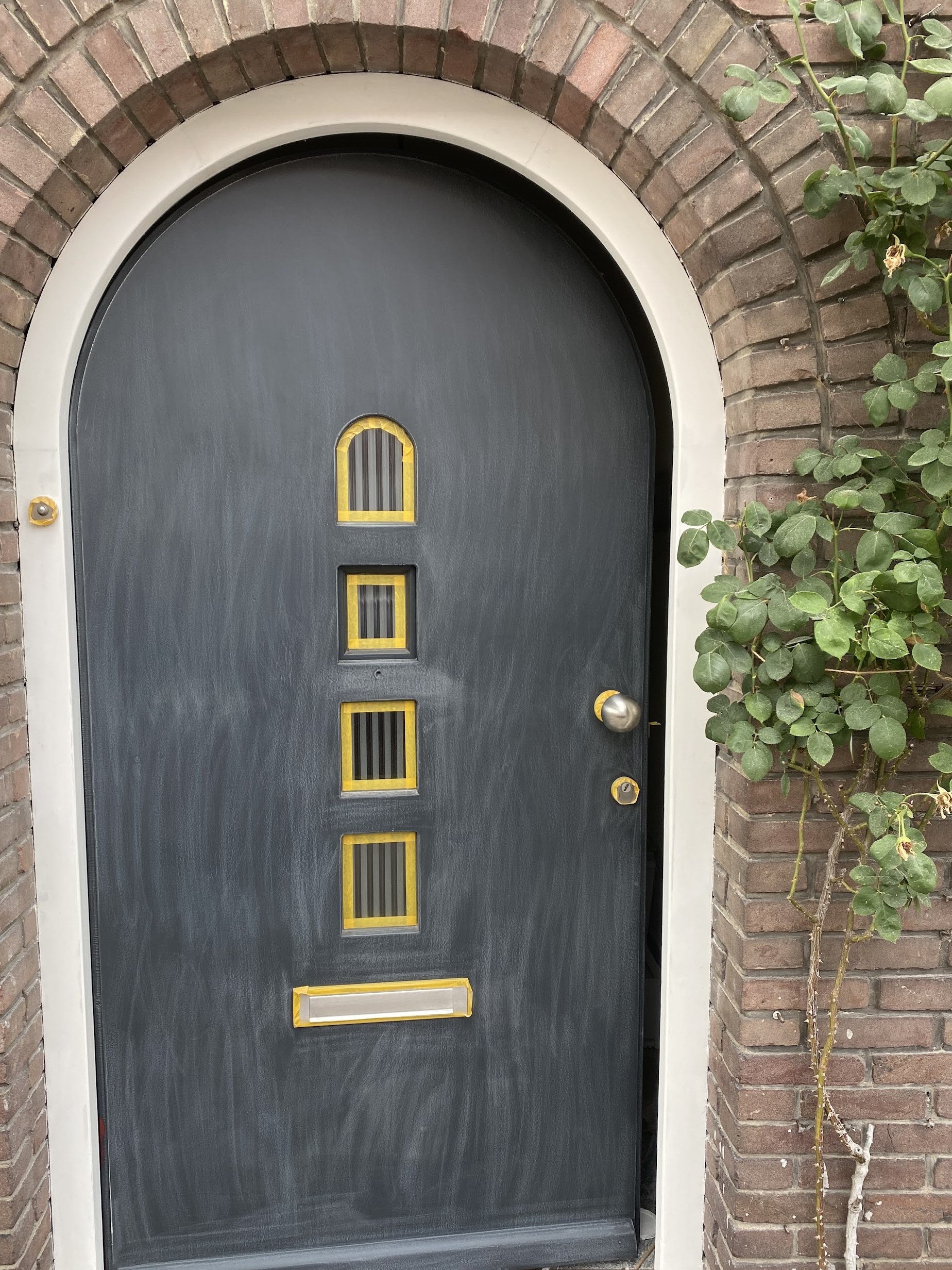 Een donkergrijs geschilderde voordeur met boog en goudkleurige roeden, in een bakstenen gevel